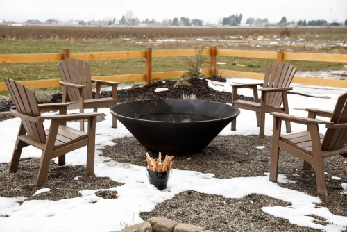 A fire pit surrounded by four wooden chairs, set in a snowy outdoor area with a wooden fence in the background.